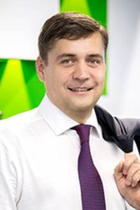 Мужчина в белой рубашке и галстуке держит пиджак на плече, стоя в помещении на фоне зеленых и белых стен.