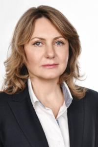 Человек с волосами до плеч, одетый в темный пиджак и белую рубашку, смотрит вперед на однотонном фоне.