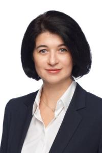 Женщина с темными волосами до плеч, в белой блузке и темном пиджаке, слегка улыбается на простом белом фоне.